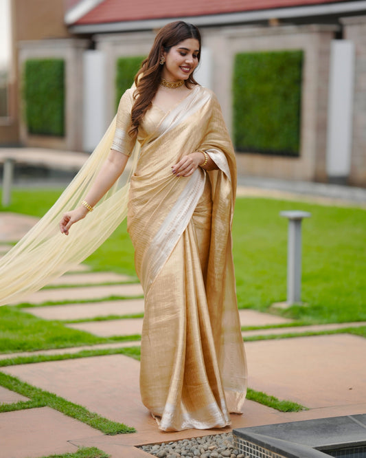 Woman in Devtha's gold saree standing outdoors with greenery and a building in the background