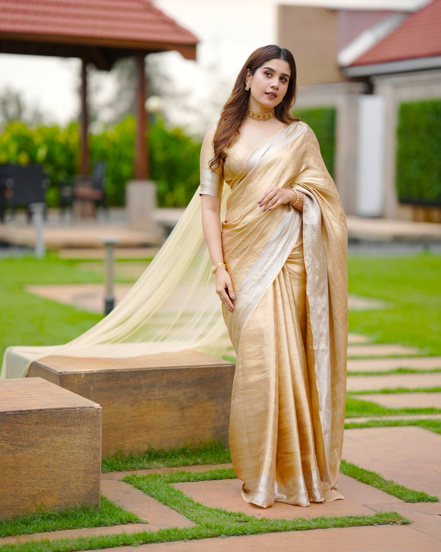 Woman in a gold saree standing outdoors on a grassy area.